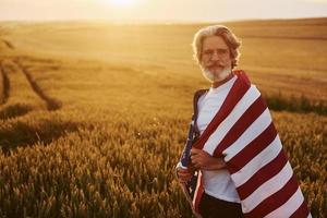 View from behind. Holding USA flag in hands. Patriotic senior stylish man with grey hair and beard on the agricultural field photo