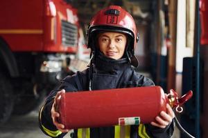 Holds extinguisher in hands. Female firefighter in protective uniform standing near truck photo