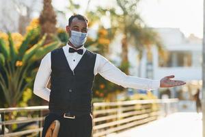 In protective mask. Black waiter in formal clothes is at his work outdoors at sunny daytime photo