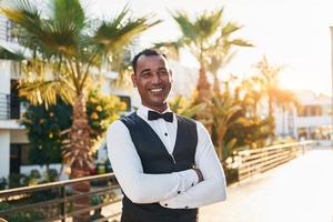 Posing for a camera. Black waiter in formal clothes is at his work outdoors at sunny daytime photo