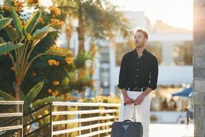 With travel bag. Young man is outdoors at sunny daytime. Concept of vacation photo