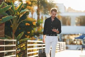 With travel bag. Young man is outdoors at sunny daytime. Concept of vacation photo