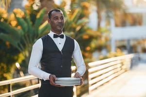 Holds empty plate. Black waiter in formal clothes is at his work outdoors at sunny daytime photo