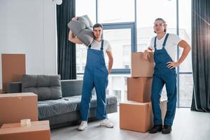 Busy day. Two young movers in blue uniform working indoors in the room photo