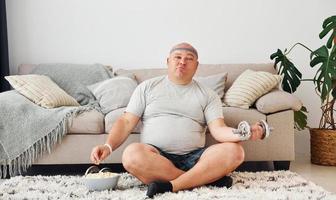 Eats and does exercises. Funny overweight man in casual clothes is indoors at home photo