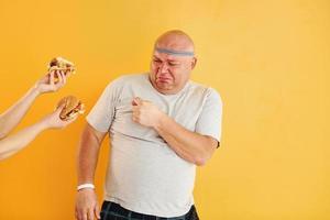 Hands with hamburgers temptates guy. Funny overweight man in sportive head tie is against yellow background photo