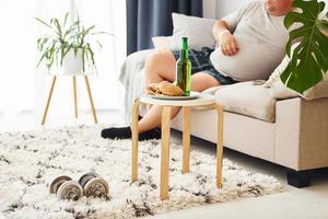 Hamburgers and beer. Man lying on the sofa. Funny overweight man in casual clothes is indoors at home photo