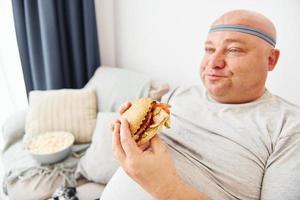 Sits on the sofa with popcorn and hamburger. Funny overweight man in casual clothes is indoors at home photo