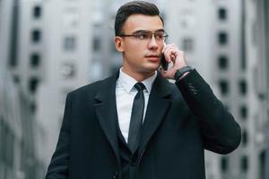 Talking by phone. Businessman in black suit and tie is outdoors in the city photo