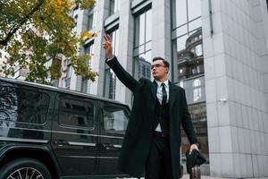 de pie contra el coche negro. hombre de negocios con traje negro y corbata está al aire libre en la ciudad foto