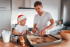 de pie junto a la mesa. padre enseñando a su pequeño hijo a preparar dulces galletas navideñas foto