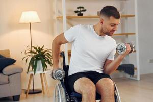 Lifting dumbbells. Disabled man in wheelchair is at home photo