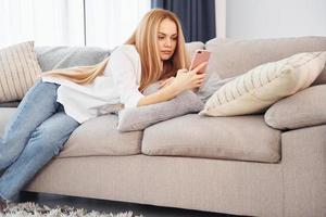 Lying down on sofa. Young woman in white shirt and jeans is at home photo