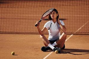 Brunette is sitting on the ground. Female tennis player is on the court at daytime photo