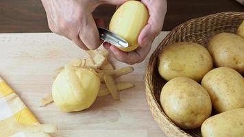 Hands peel potato, peelings on wooden cutting board.