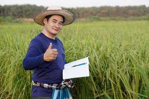 Handsome Asian man farmer is at paddy field, wear hat, blue shirt, holds notebook paper, inspects growth and disease of plants. Concept, Agriculture research and study to develop crops. photo