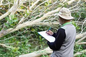 Asian man botanist is at forest to survey botanical plants, holds paper clipboard. Concept , Survey ,research botanical plants. Forest and environment conservation. photo