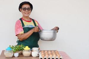 Happy Asian senior woman is cooking, wear chef cap and apron, holds pot, smile. Concept, Cooking for family. Thai kitchen lifestyle. Elderly activity. photo