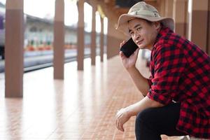 Asian man traveler is waiting someone at platform of train station, holds mobilephone to talk on the phone. Concept, travel alone, transportation, communication or connection. photo