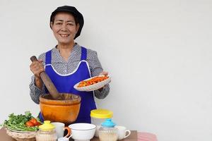 Happy Asian senior woman is cooking, wear chef cap and apron, holds pestle, mortar and plate of chillies. Concept, Cooking for family. Thai kitchen lifestyle. Elderly activity. photo