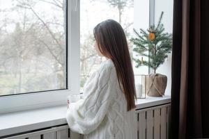 Woman in warm white winter sweater standing next to the window at home at christmas eve holding cup with marshmallows, fir tree behind photo
