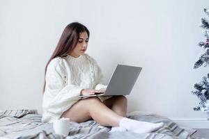 Woman in warm white winter sweater lying in bed at home using laptop photo