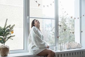 mujer con suéter de invierno blanco cálido sentada en el alféizar de la ventana en casa en la víspera de navidad sosteniendo una taza con malvaviscos foto