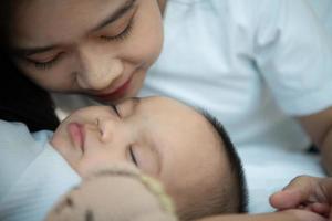 joven madre abrazando a su hijo recién nacido para arrullar al bebé a dormir en el dormitorio blanco foto