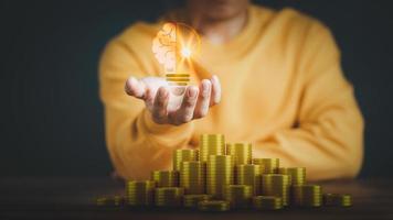 Man holding a glowing abstract light bulb with pile money cions on a wooden table, business problem solving ideas and creative marketing ideas Inventing and learning new things. photo