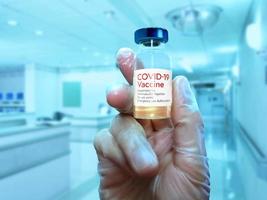 A close-up hand with a glove holding COVID-19 vaccine over a blurred hospital background photo