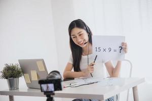 una joven y hermosa profesora asiática enseña matemáticas en línea y mira una laptop para chatear con un estudiante frente a la cámara durante la cuarentena y el trabajo desde casa.