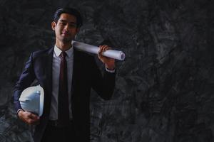 Young business engineering holding white helmet and blue print on dark background, smart and confident worker. photo