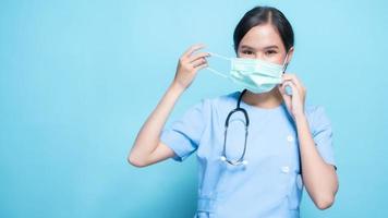 retrato de una joven enfermera asiática hermosa con uniforme azul y con una máscara quirúrgica para protegerse de la enfermedad de covid19 o coronavirus aislada en el fondo azul del estudio.