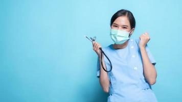 Asian Thai nurse or doctor wearing mask and holding stethoscope with big smiled isolated in studio on blue background photo