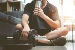 hombre bebiendo proteína o bebida de recuperación sentado en el suelo de un gimnasio después de un entrenamiento. hacer ejercicio y hacer ejercicio en casa. foto