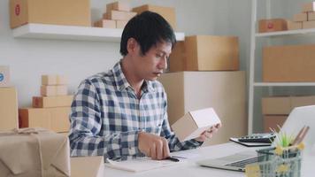 Asian male business owner is preparing online order for delivery via courier company. Small business owners are checking notes and jotting down warehouse details and collecting them for delivery. video