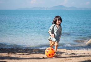 niño asiático feliz jugando fútbol descalzo en la playa con una pelota roja y naranja durante el día, niño divirtiéndose al aire libre. concepto de vacaciones de verano. foto