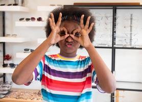 el retrato de una niña africana brillantemente sonriente pone su dedo en un semicírculo que simboliza anteojos mientras selecciona un marco en una tienda de óptica. foto