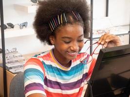 niña africana en la tienda óptica mirando en el espejo para elegir un nuevo marco de anteojos. concepto de salud, medicina, personas, vista y tecnología foto