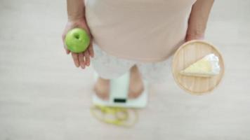 concepto de dieta. las mujeres están eligiendo los alimentos adecuados para una buena salud. las mujeres eligen entre pastel de chocolate blanco y manzanas verdes durante la medición del peso en básculas digitales. video