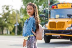 niña de pie junto a un gran autobús escolar con su mochila rosa. foto