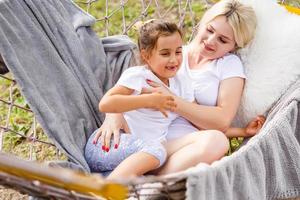 Beautiful young mother and daughter laying down and relaxing together on a hammock during a sunny summer day on holiday home garden. Family relaxing outdoors, healthy and wellness lifestyle. photo