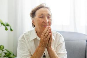 Faithful old senior grandmother pray with hope faith holding hands together sit on sofa alone at home, religious elderly woman believer christian with eyes closed say gratitude worship prayer concept photo