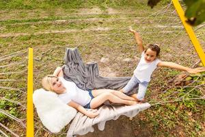 Beautiful young mother and daughter laying down and relaxing together on a hammock during a sunny summer day on holiday home garden. Family relaxing outdoors, healthy and wellness lifestyle. photo