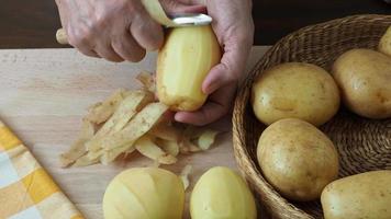 Hands peel potato, peelings on wooden cutting board.