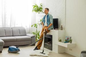 Caucasian handsome serious young man vacuuming cleaning modern apartment, clean up, housekeeper, guy doing domestic tasks and cleans living room using vacuum cleaner, householding photo