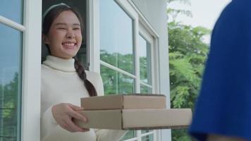 una mujer asiática feliz y sonriente recibe una caja de comida del servicio de mensajería en la casa principal. repartidor en uniformes entregar express. compras en línea, contenedores de papel, comida para llevar, cartero, servicio de entrega video