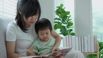 contento asiatico madre rilassare e leggere libro con bambino tempo insieme a casa. genitore sedersi su divano con figlia e lettura un' storia. imparare sviluppo, assistenza all'infanzia, ridendo, formazione scolastica, narrativa, la pratica. video