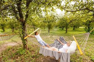 Beautiful young mother and daughter laying down and relaxing together on a hammock during a sunny summer day on holiday home garden. Family relaxing outdoors, healthy and wellness lifestyle. photo