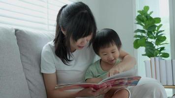 heureuse mère asiatique se détendre et lire un livre avec bébé temps ensemble à la maison. un parent s'assoit sur un canapé avec sa fille et lit une histoire. apprendre le développement, la garde d'enfants, le rire, l'éducation, la narration, la pratique. video